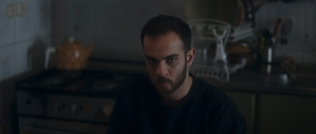 A man sitting on the chair, wearing a black sweater and looking away. A frame from the feature film Withered Pomegranates. Behrad Selahvarzi