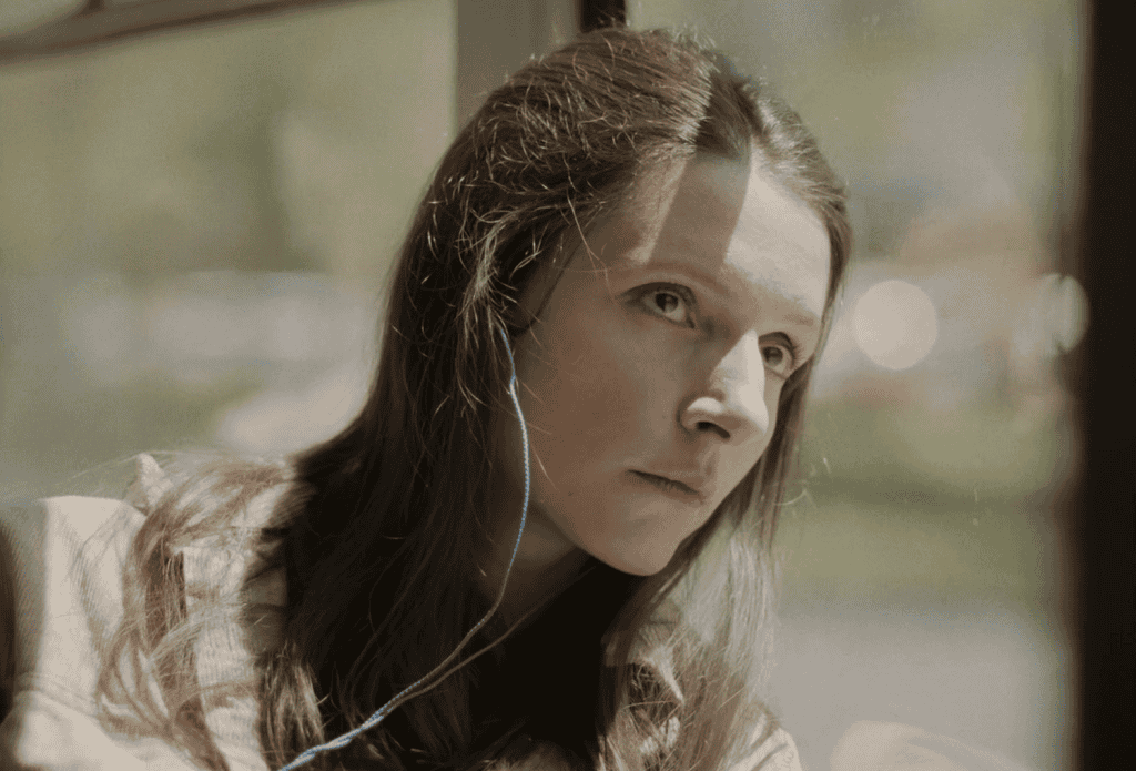 Sara sitting alone on a bus with her head against the window, listening to music with headphones on.