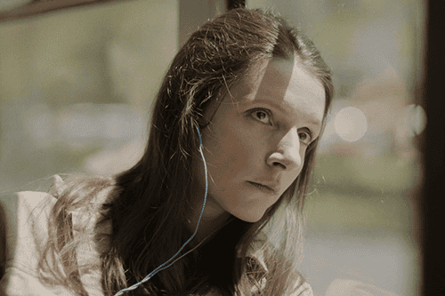 Sara sitting alone on a bus with her head against the window, listening to music with headphones on.